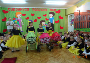 Na scenie 4 dziewczynki w czarnych body i żółtych spódnicach stoją z trzema wózkami. Po bokach siedzą chłopcy ubrani na galowo.